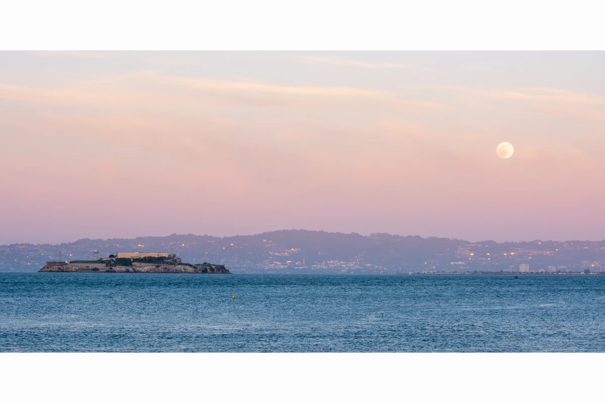 Alcatraz under a pink sky at sunset, with the moon rising over the East Bay hills.